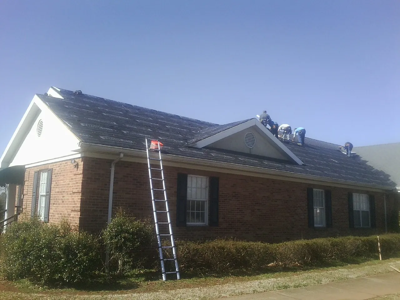 Crew tearing shingles off a brick ranch roof with a ladder set — Emergency Roof Repair work by our crew in Dousman