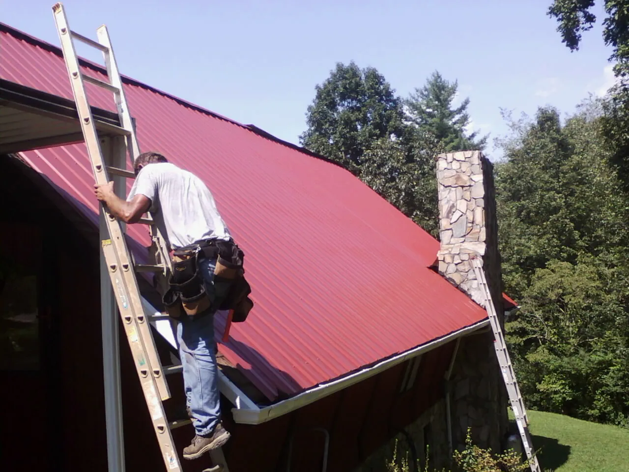 Roofer on a ladder beside a red metal roof and stone chimney — Soffit & Fascia Repair by Roofing Brothers in Dousman
